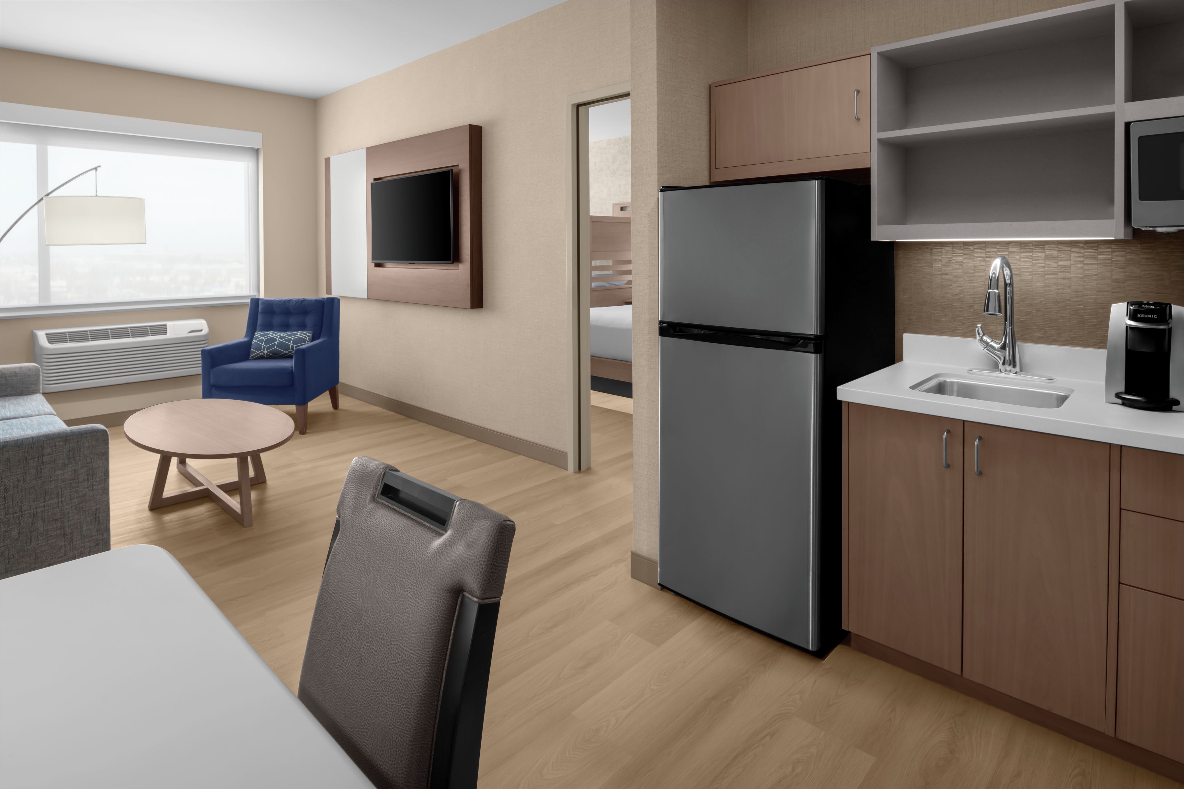 A modern hotel room with a kitchenette, a seating area with a round table and chairs, a flat-screen TV mounted on the wall, and a large window providing a view.