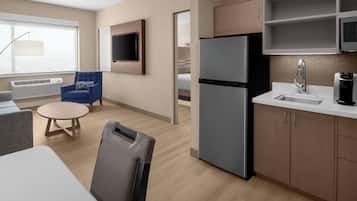 A modern hotel room with a kitchenette, a seating area with a round table and chairs, a flat-screen TV mounted on the wall, and a large window providing a view.