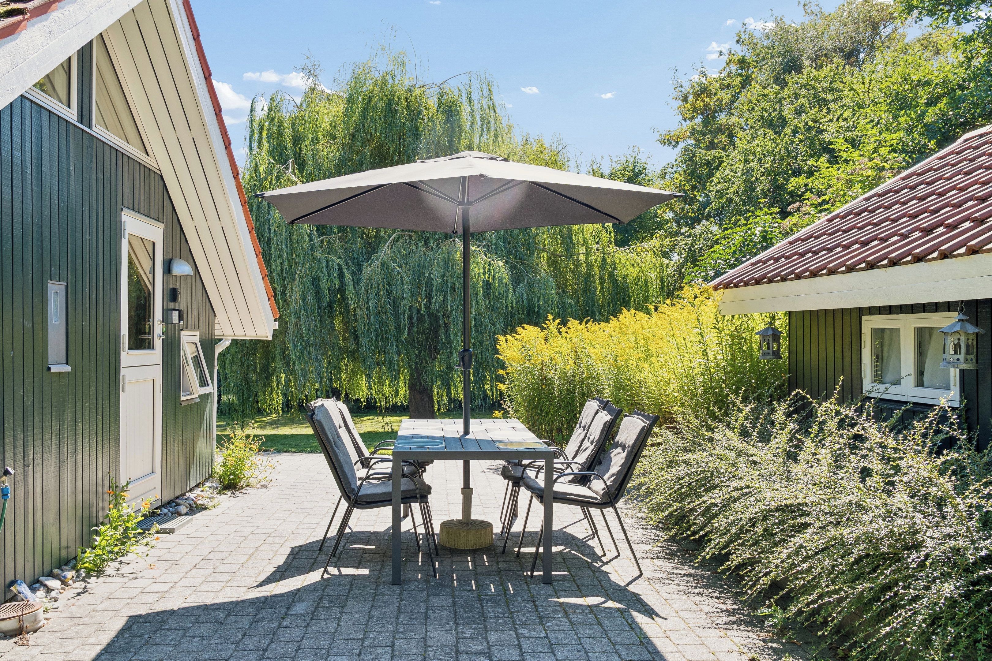 Een terras buiten met een tafel en stoelen, een grote parasol, en een tuin met diverse planten.