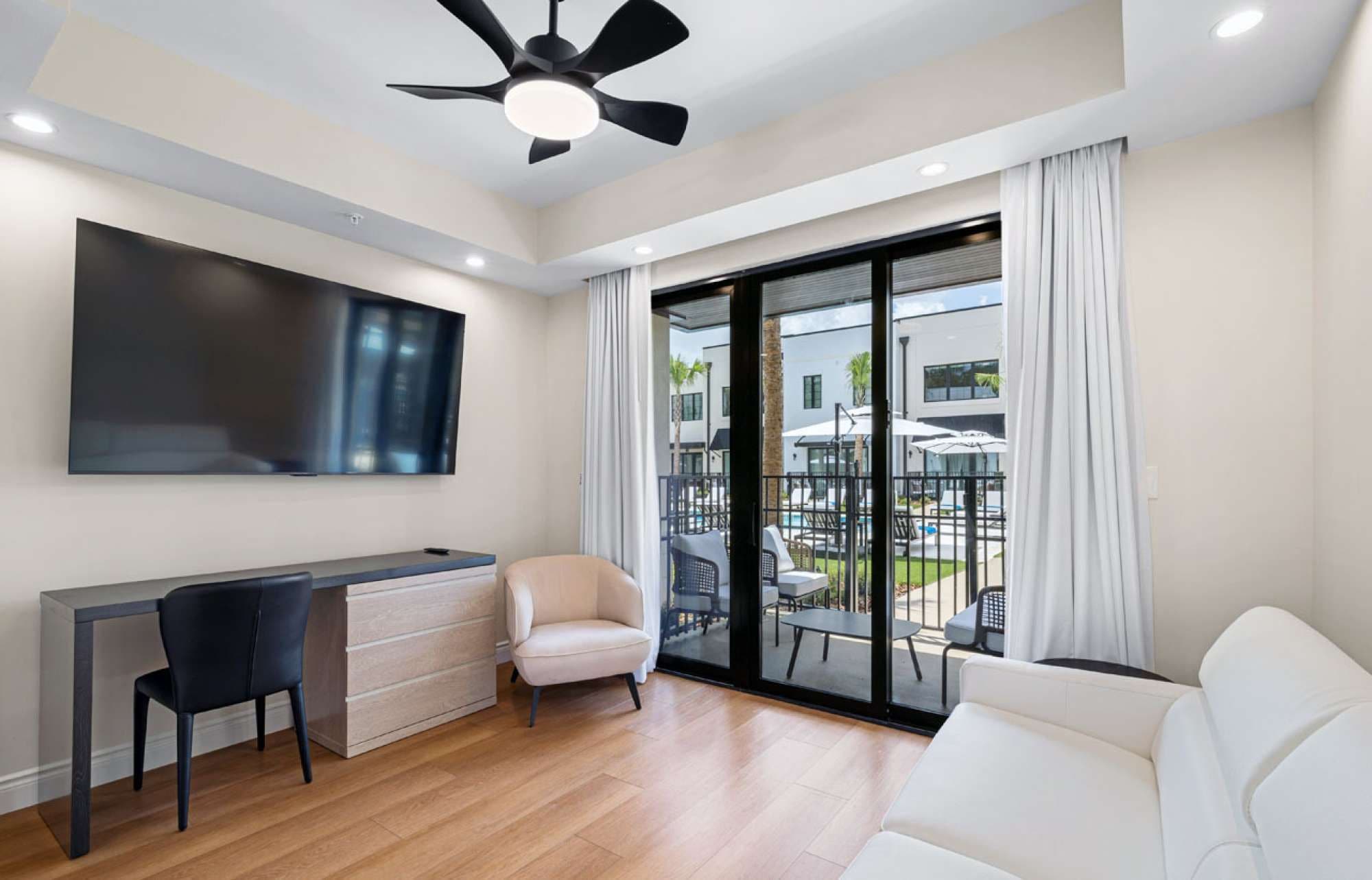 A modern living room with a flat-screen TV, a desk, a chair, a sofa, and a balcony with outdoor furniture.