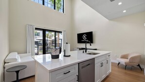 Eine moderne Küche mit Flachbildfernseher, einem Essbereich mit weißem Tisch und Stühlen sowie Blick ins Freie durch ein großes Fenster.