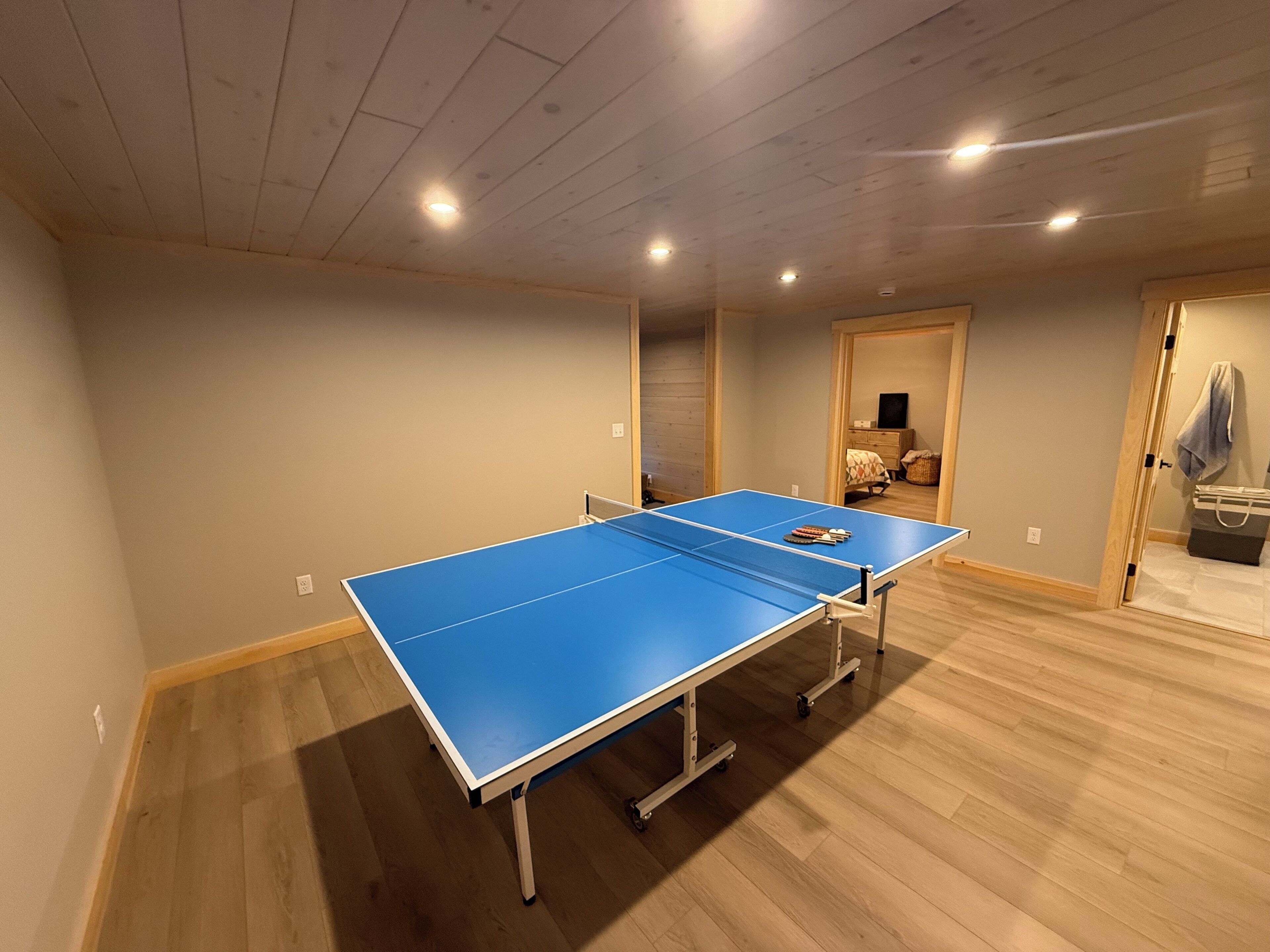 A room with a ping pong table, wooden flooring, and a door leading to another room.
