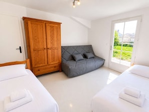 A bedroom with two beds, a grey sofa, a wooden wardrobe, and a large window with a view of greenery. - Lovely, fully equipped holiday home (LA COUARDE SUR MER)