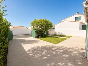 A courtyard with a paved walkway, a small lawn, and a building with a white facade and green shutters. - Lovely, fully equipped holiday home (LA COUARDE SUR MER)