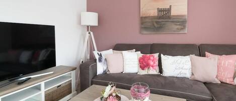 A modern living room with a flat-screen TV, a sofa with decorative pillows, a coffee table with a candle and a small plant, and a painting on the wall.