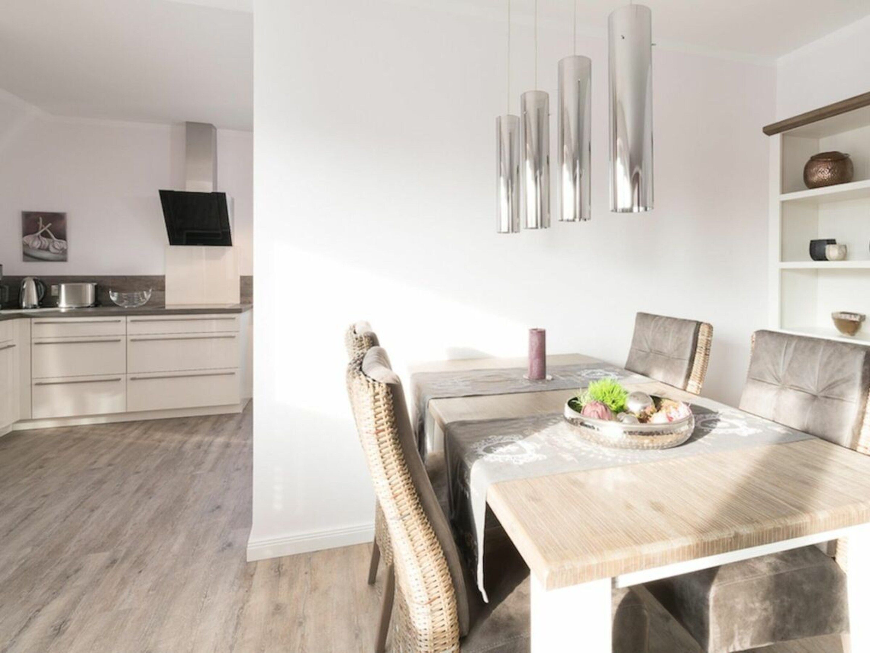 Un comedor moderno con una mesa de madera y sillas de mimbre, una lámpara colgante y una cocina al fondo.