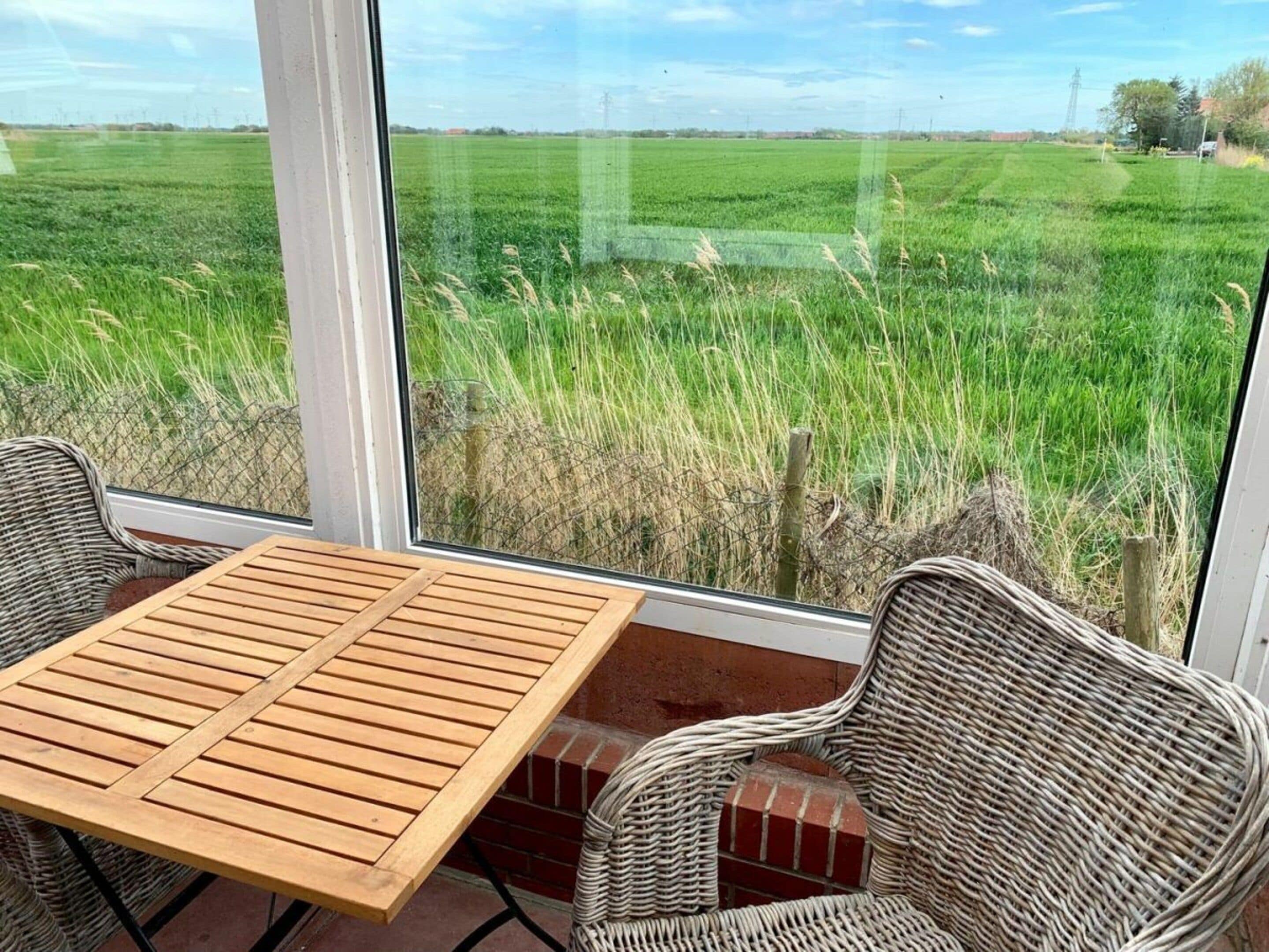 A room with a large window overlooking a green field, a wooden table, and wicker chairs.