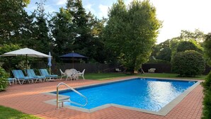 Ein rechteckiger Schwimmteich mit einer Metallleiter, umgeben von einer Ziegelterrasse. Liegestühle und ein Tisch mit Sonnenschirmen sind vorhanden.