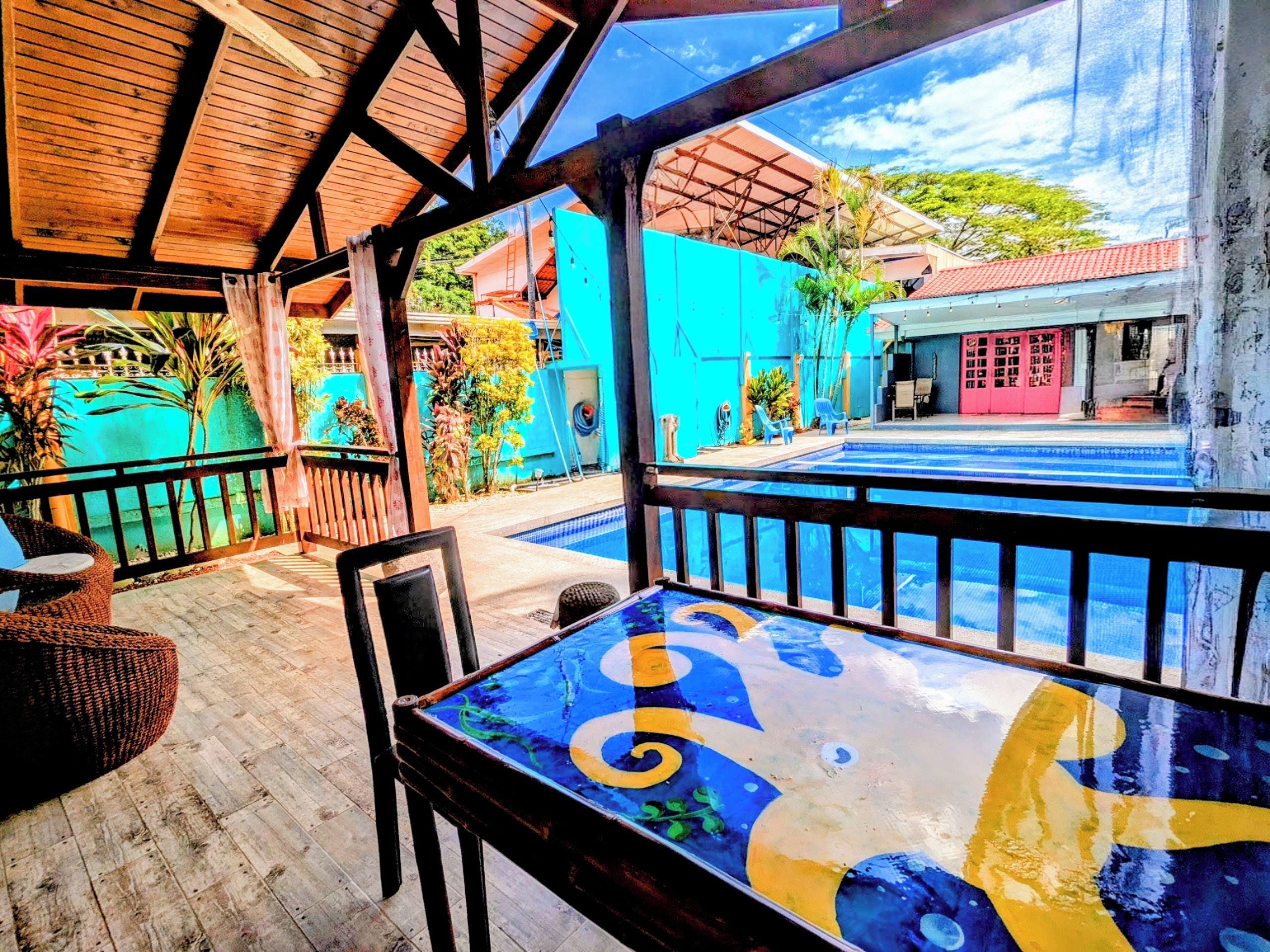 Un espace extérieur aménagé, abrité et doté de sièges, avec une piscine, un plancher en bois et une vue sur un bâtiment bleu.