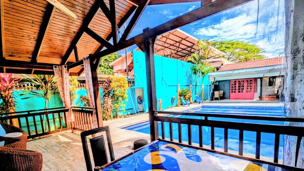 A covered outdoor seating area with a pool, wooden flooring, and a view of a blue building. - Tropical Paradise w/ 2 Sapacious Cabinas AC & Pool (Playa Herradura)