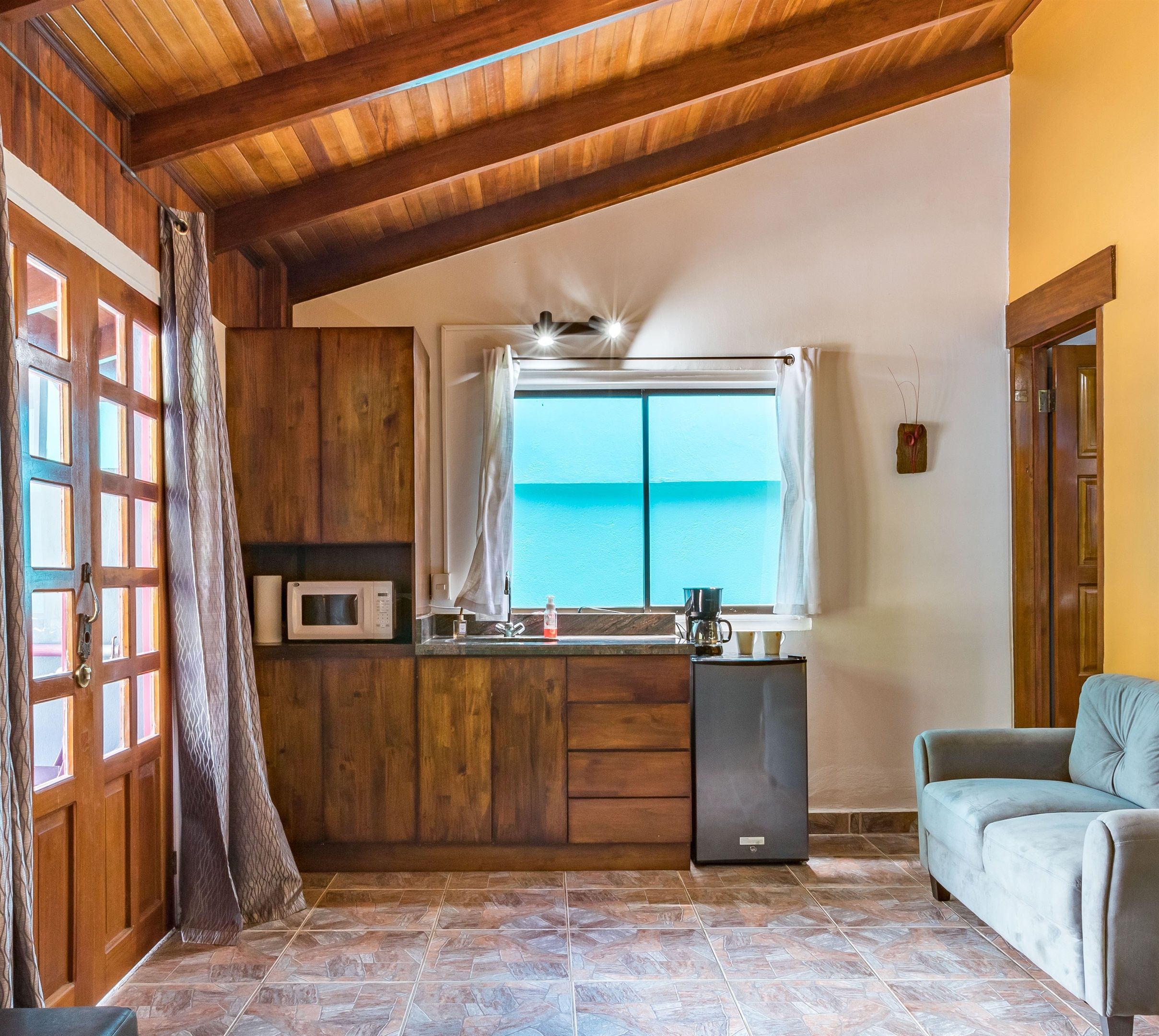 Cocina compacta con armarios de madera, microondas y refrigerador. Hay una ventana con vista al mar, y un sofá en la sala.