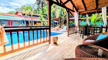 Una terraza de madera con piscina, sillas de descanso y vistas a un paisaje tropical.