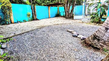 Un patio de grava con valla azul, árboles y un edificio blanco al fondo.