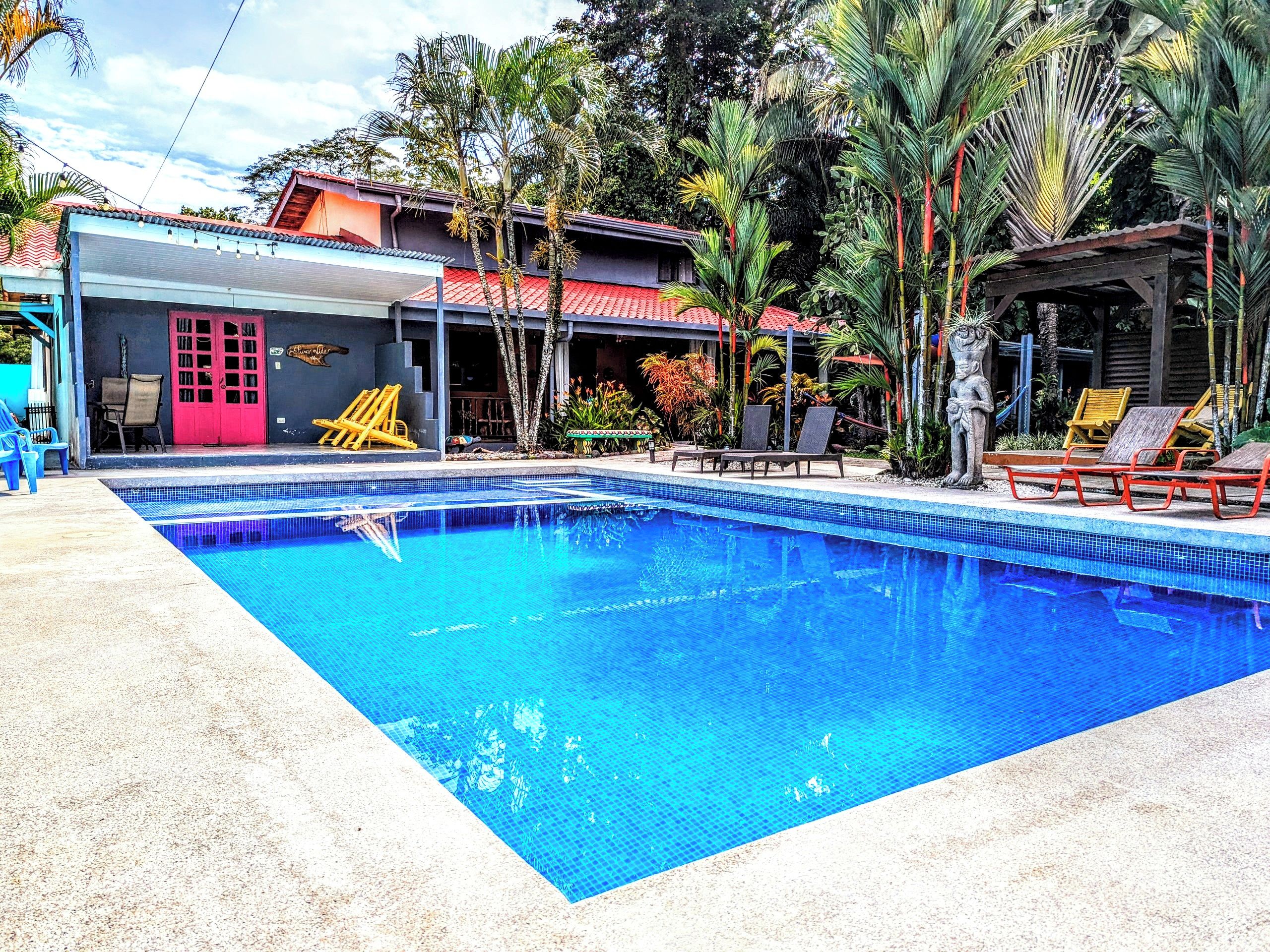 Una piscina con lettini e un edificio con una porta rossa e un tetto in tegole.