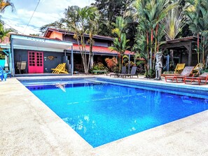 A swimming pool with lounge chairs and a building with a red door and a tiled roof.