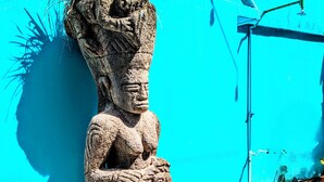A stone statue of a figure with a plant on its head, standing against a bright blue wall.