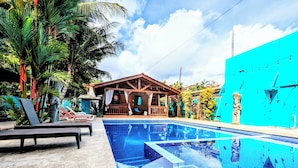 A swimming pool with a wooden deck, lounge chairs, and a building with a porch. - Stylish & Cozy Private Rooms in Tropical Paradise (Playa Herradura)