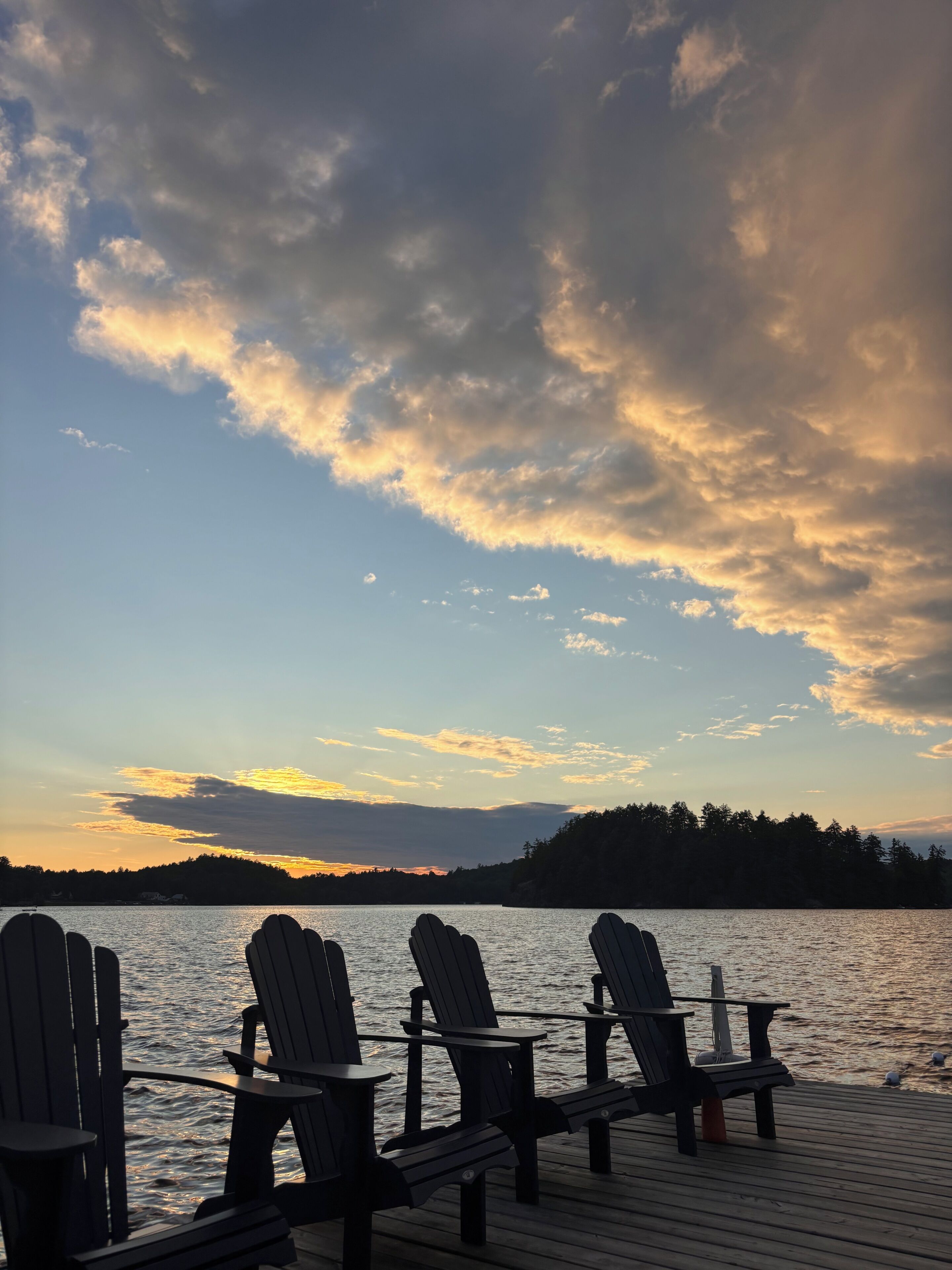 Eine Holzterrasse mit mehreren Adirondack-Stühlen, die auf ein Gewässer ausgerichtet sind, mit einem bewaldeten Hügel im Hintergrund.