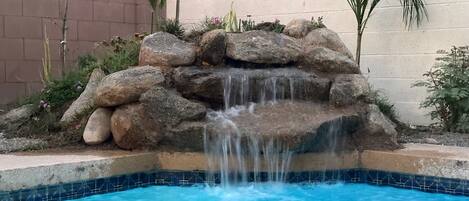 Piscina com cachoeira artificial, rodeada por pedras e plantas.