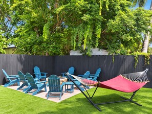 Un jardin avec un espace détente comprenant des fauteuils Adirondack bleus, un hamac rouge et un foyer.