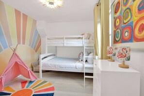 Ein Kinderstockbett mit weißem Gestell, ein kleiner Schreibtisch mit einer Flamingolampe und ein farbenfrohes Wandbild.