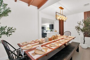 A dining area with a long wooden table set for a meal, a modern kitchen in the background, and a decorative light fixture above the table.