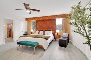 A modern bedroom with a wooden headboard, a bed with white bedding, a teal bench, a desk with a laptop, and a ceiling fan.
