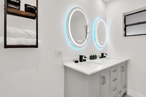 Une salle de bain moderne avec deux miroirs éclairés, un lavabo blanc et un meuble-lavabo avec des tiroirs.