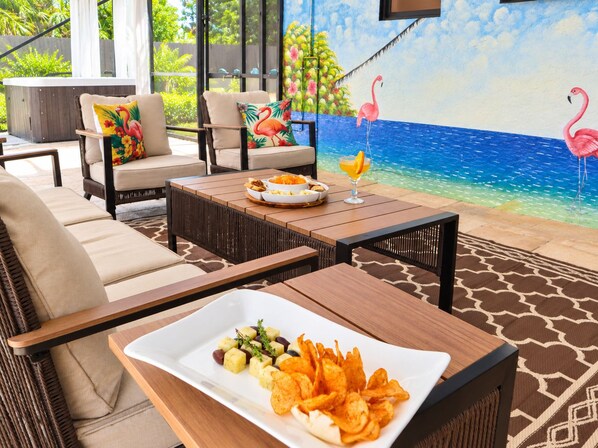 An outdoor seating area with wicker furniture, a dining table with a plate of snacks, and a mural of a beach scene with flamingos.