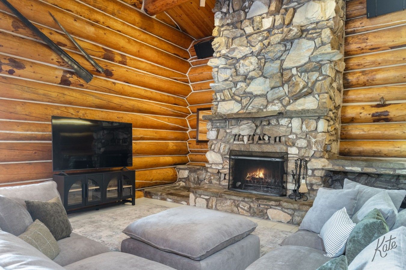 Interior de una cabaña de troncos con chimenea de piedra, televisor y sofá.