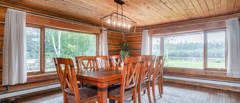A wooden interior with a dining table and chairs, large windows, and a hanging light fixture.