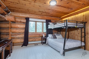 A log cabin bedroom with a bunk bed, a bedside table, a window with curtains, and a small desk. - Chena Riverside Lodge (Fairbanks)