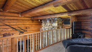 A log cabin interior with a stone wall, a wooden railing, a fireplace, and a television. - Chena Riverside Lodge (Fairbanks)