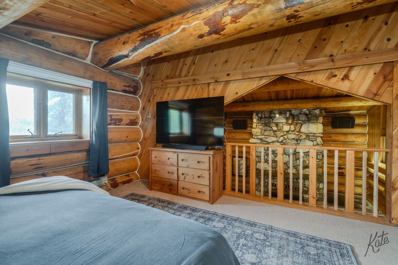 A log cabin bedroom with a bed, a dresser, a television, and a stone wall.