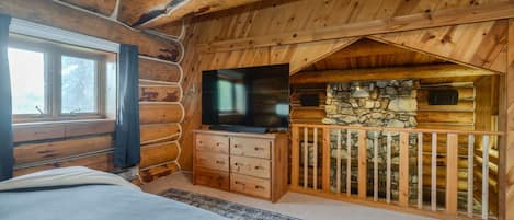 A log cabin bedroom with a bed, a dresser, a television, and a stone wall.