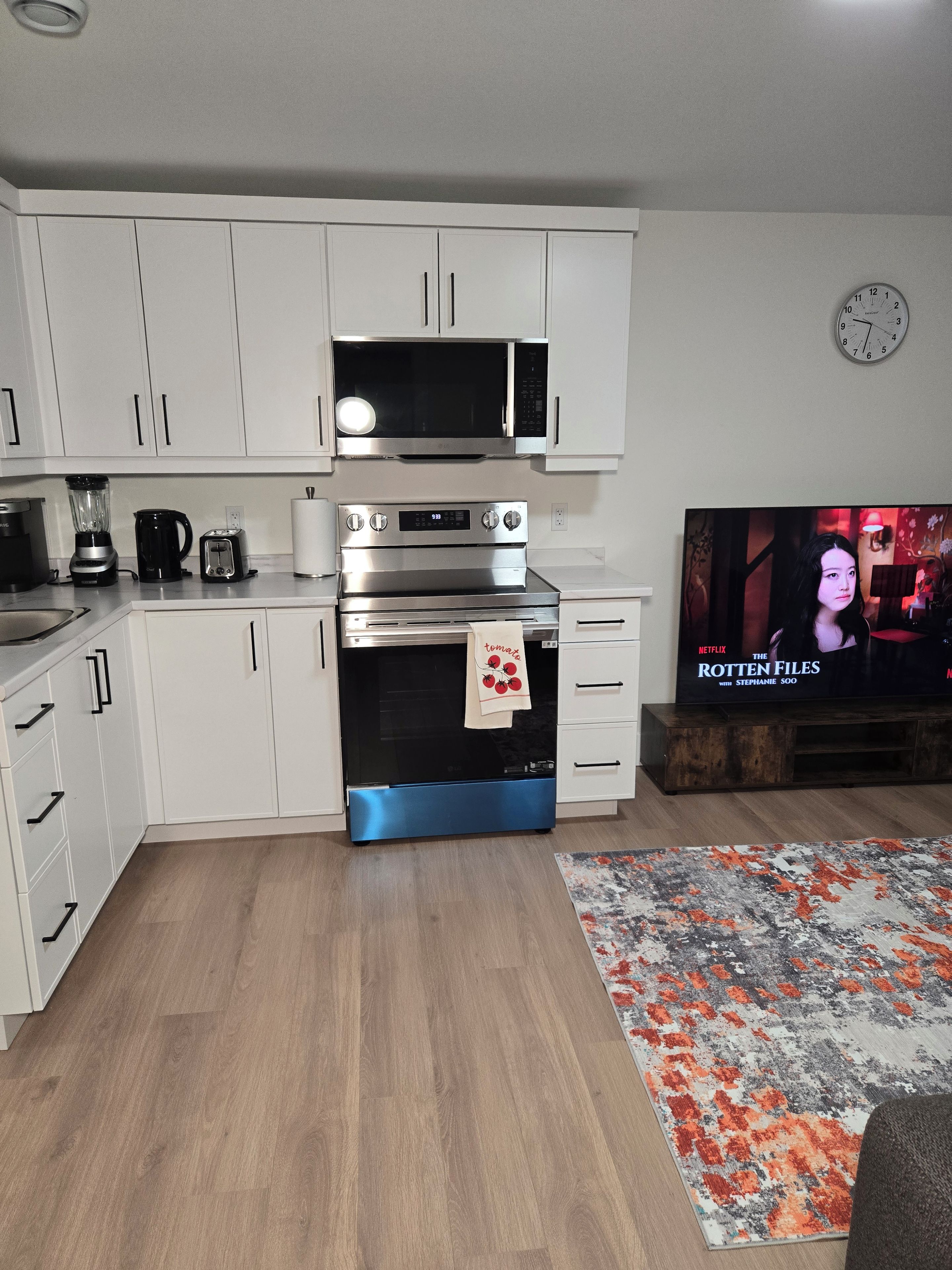 A modern kitchen with white cabinets, a microwave, a toaster, and a stove. There is a wooden floor and a rug. A television displaying 'The Rotten Files' is visible.