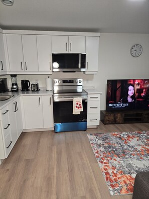 Eine moderne Küche mit weißen Schränken, einer Mikrowelle, einem Toaster und einem Herd. Ein Holzboden und ein Teppich sind vorhanden. Ein Fernseher zeigt die Sendung 'The Rotten Files'.