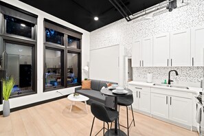 A modern kitchen with white cabinetry, a dining area with a grey sofa and black chairs, and large windows with reflections of the outside. - Your Downtown Base | Walk to Petco Park & Gaslamp! (San Diego)