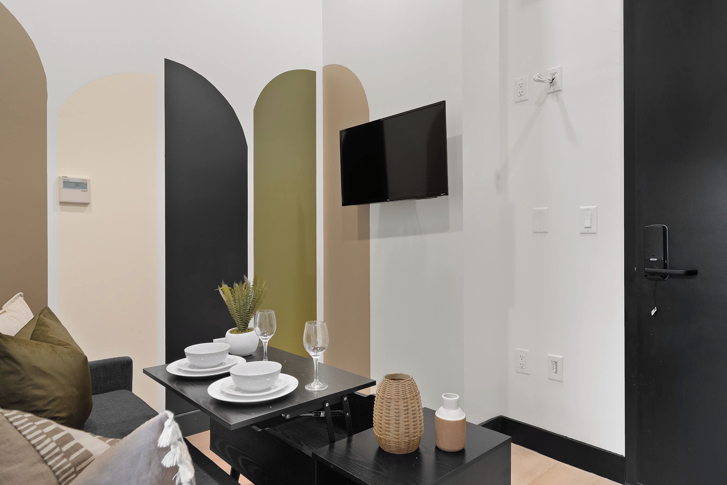 A modern living room with a black sofa, a dining table set with plates and glasses, a flat-screen TV mounted on the wall, and a door with a handle.