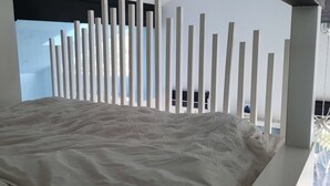 Ein Bett mit einer strukturierten weißen Bettdecke, eine weiße Wand mit vertikalen Lamellen und ein Blick ins Freie durch ein Fenster.