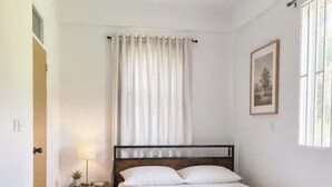 Quarto com uma cama de solteiro, mesa de cabeceira com abajur, janela com cortinas e um quadro na parede.