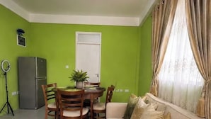 Sala de estar com sofá, mesa de jantar com cadeiras, uma geladeira pequena e uma luminária circular.