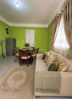 Una sala de estar con sofá, una mesa de comedor con sillas, un refrigerador pequeño y una luz de aro.