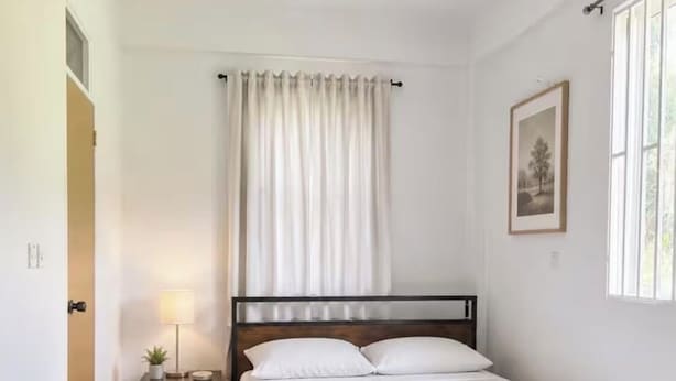 Quarto com uma cama de solteiro, mesa de cabeceira com abajur, janela com cortinas e um quadro na parede.
