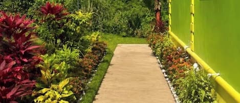 A pathway leading to a building with a green exterior, surrounded by lush vegetation and a mountainous backdrop.