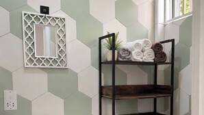 Um banheiro com parede de azulejos hexagonais, um móvel de madeira com pia, vaso sanitário e prateleira de metal com toalhas.