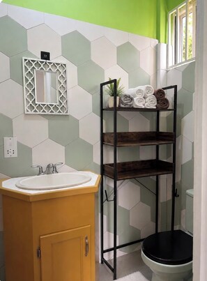 Un baño con pared de azulejos hexagonales, un mueble de tocador de madera con un lavamanos, un inodoro y un estante metálico con toallas.