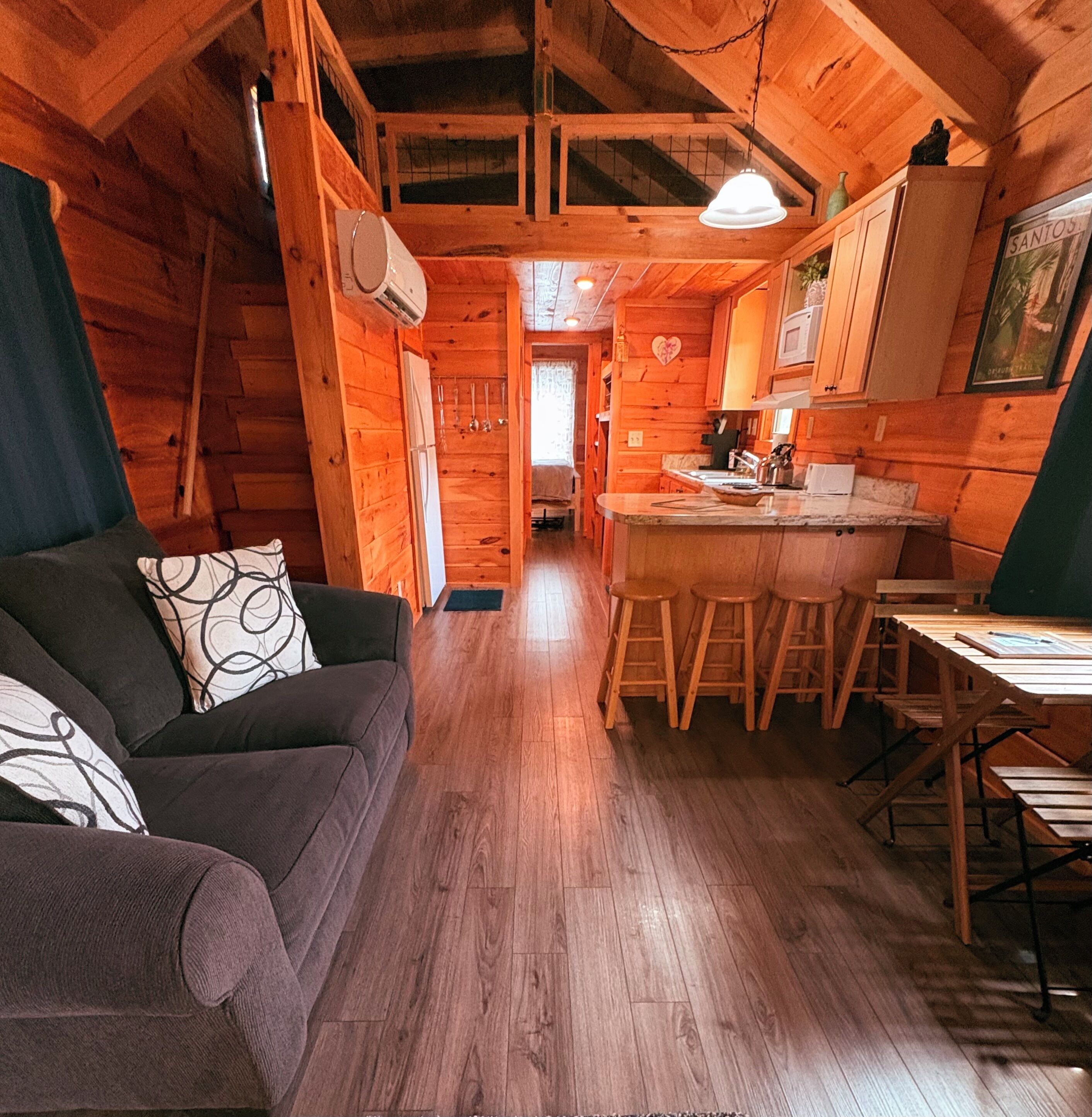 Interior acollidor d'una cabana amb sòl de fusta, un sofà amb coixins estampats, zona de menjador amb tamborets i cuina amb armaris de fusta.