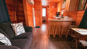 Interior acogedor de una cabaña con piso de madera, un sofá con cojines estampados, una zona de comedor con taburetes y una cocina con armarios de madera.