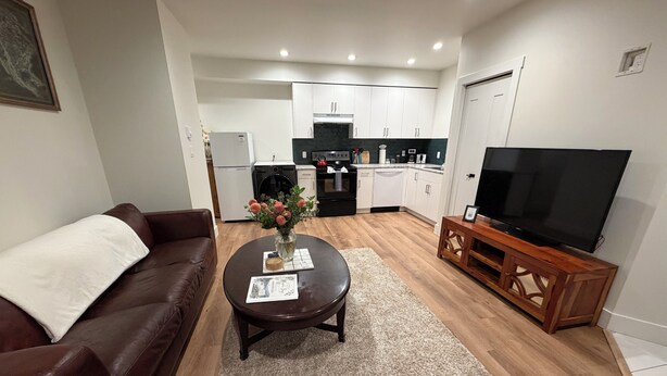 A living room with a brown leather sofa, a coffee table with a vase, a flat-screen TV on a wooden stand, and a kitchen in the background. - Cozy Two-Storey Suite in Maple Bay (Duncan)